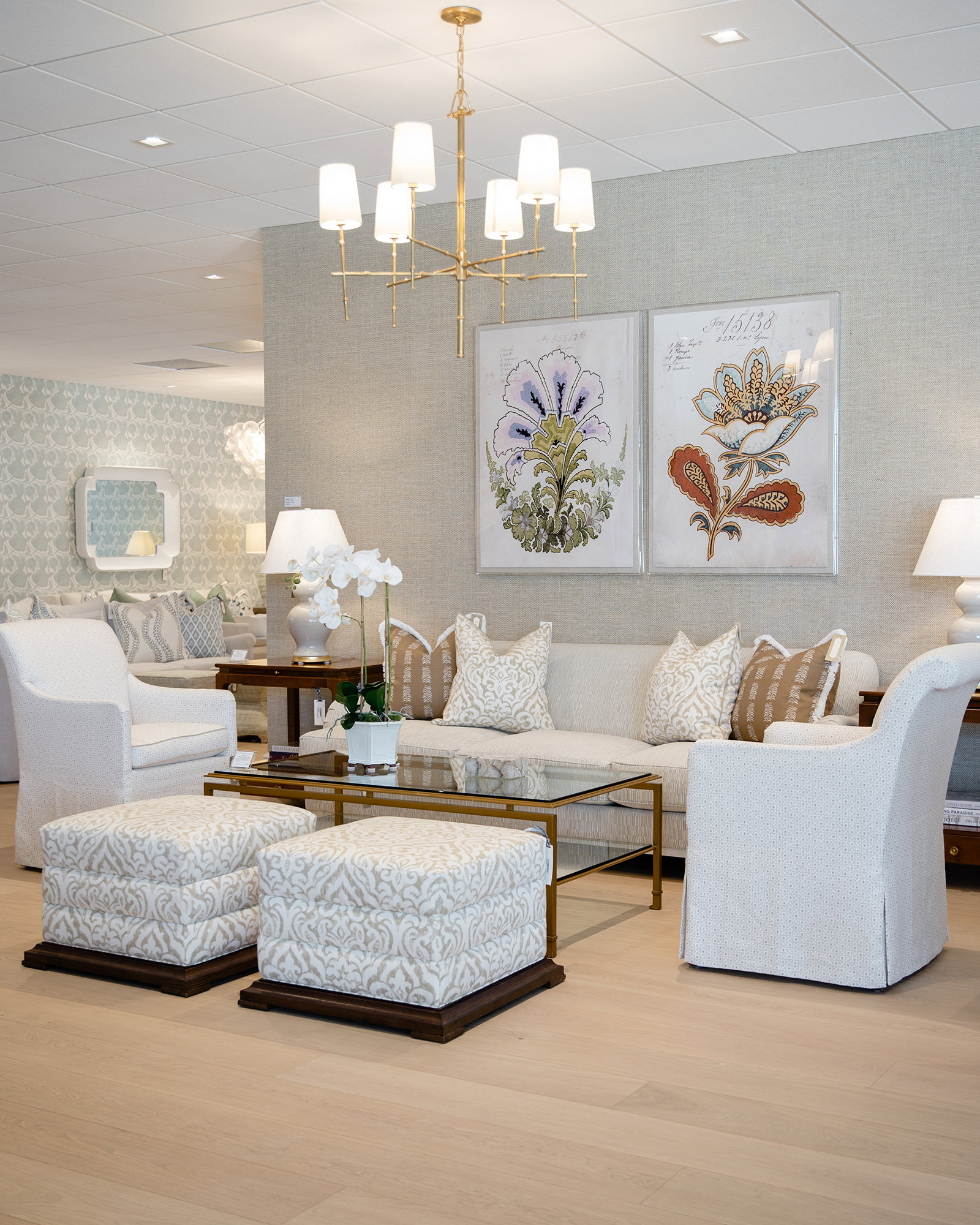 image of neutral showroom living space with white and brown upholstered stools and pillows on white sofa and chairs and botanical prints on wall