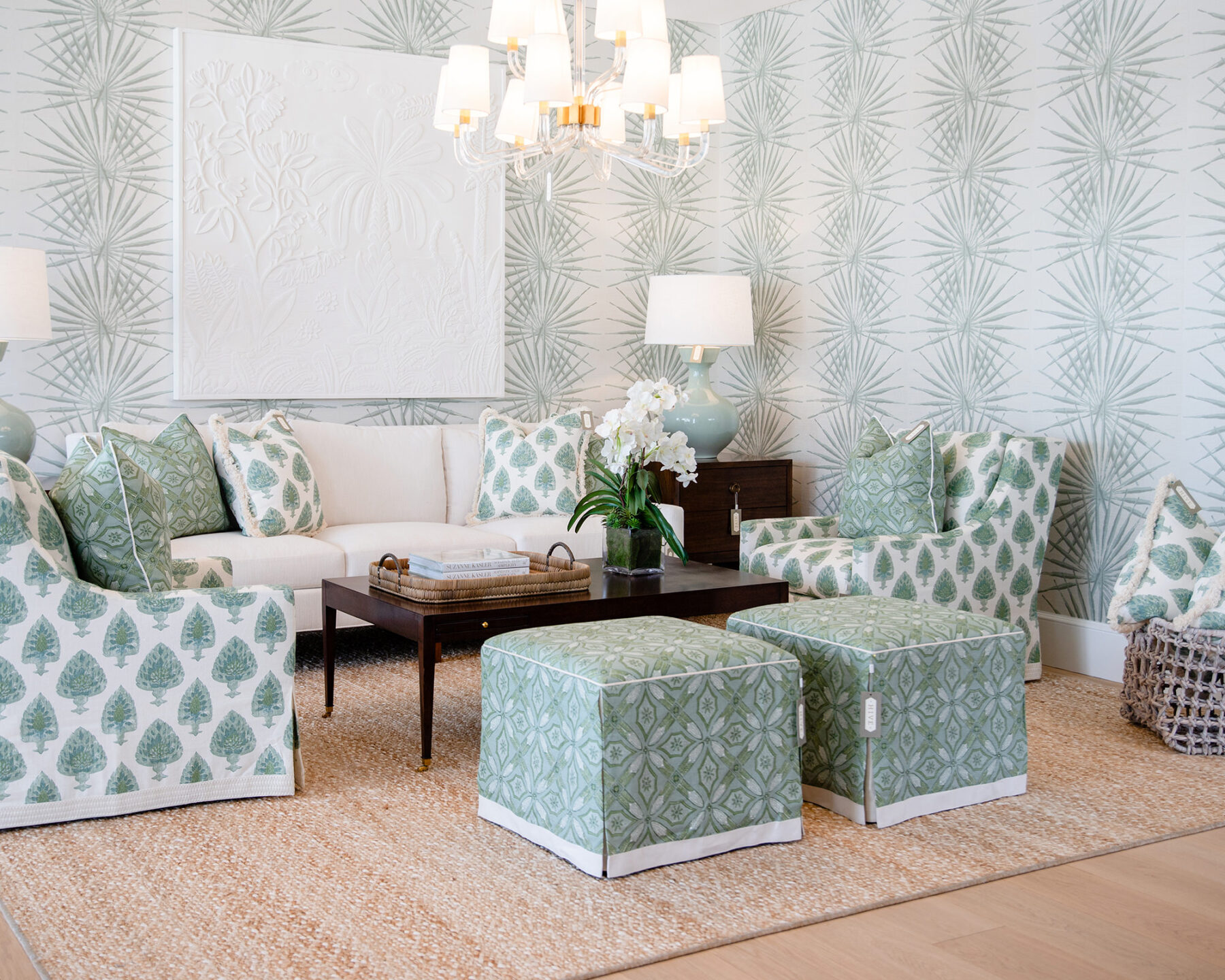 image of showroom wallpapered living area with mint green and white patterned upholstered chairs with matching pillows on a white sofa, dark walnut cocktail table and a pair of upholstered stools