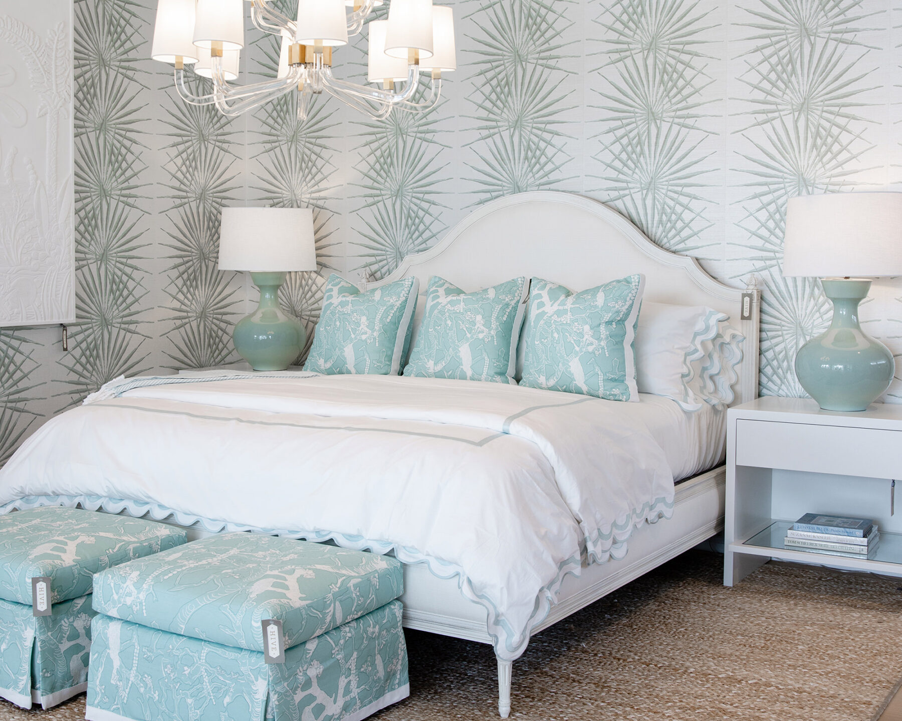 image of showroom bedroom featuring a white and wood upholstered bed, mint green patterned pillows and matching stools, a mint palm leaf wallpaper and mint table lamps atop white nightstands