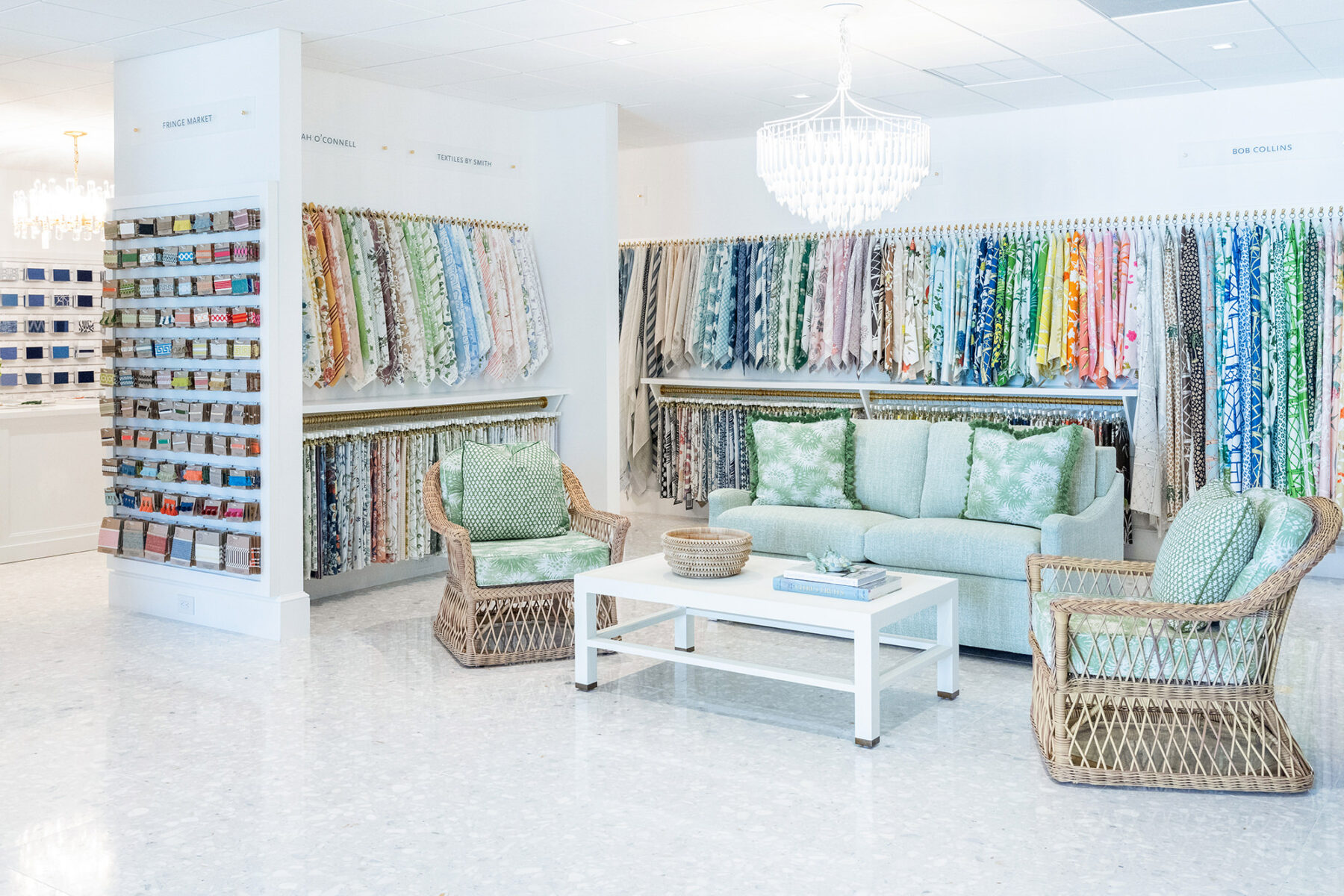 image of trade showroom fabric wall with sitting area in font featuring a free sofa, string upholstered chairs with matching upholstered green pillows and a white cocktail table