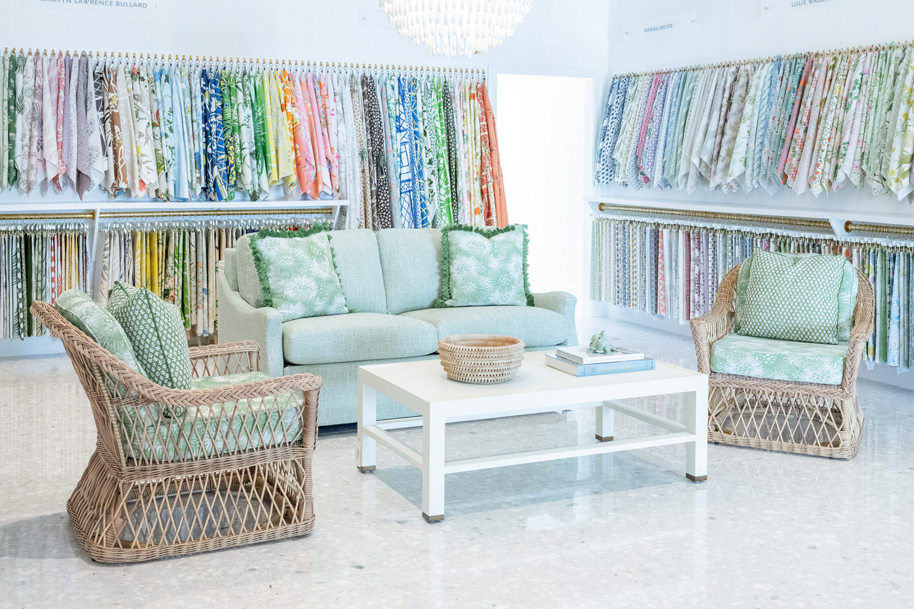 image of trade showroom fabric wall with sitting area in font featuring a free sofa, string upholstered chairs with matching upholstered green pillows and a white cocktail table